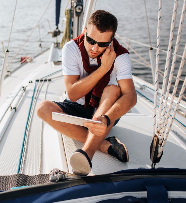 busy young man sits board yacht talks phone also he holds looks tablet young man sits with legs crossed he wears sunglasses